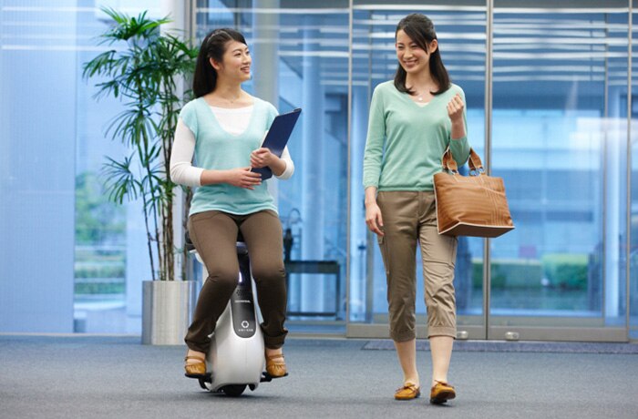 A woman walks beside a woman on a Honda UNI-CUB